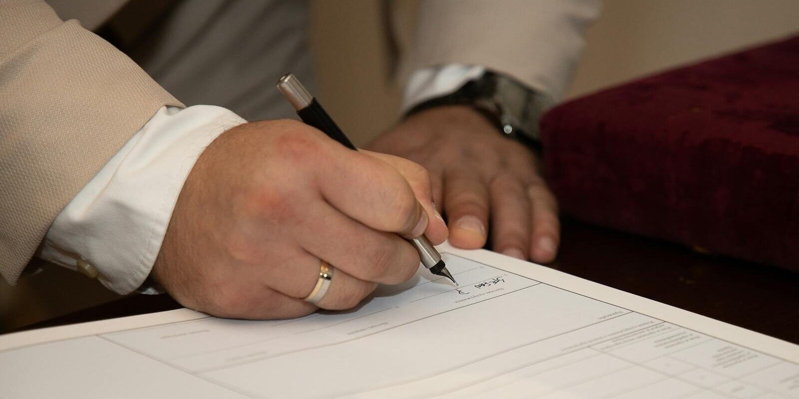 Photo by Boris Hamer A close-up of a man signing a document, showcasing a wedding ring and pen.