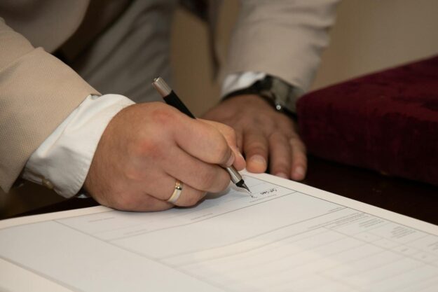 Photo by Boris Hamer A close-up of a man signing a document, showcasing a wedding ring and pen.